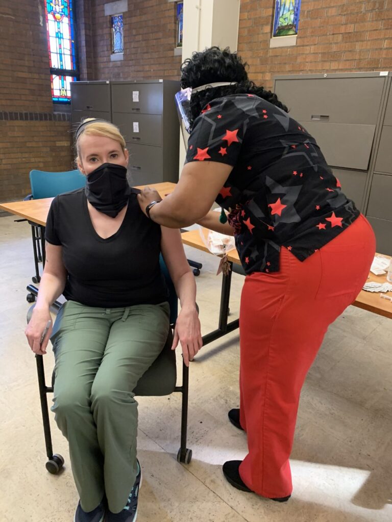 Woman seated in chair wearing surgical mask receives a shot from healthcare provider