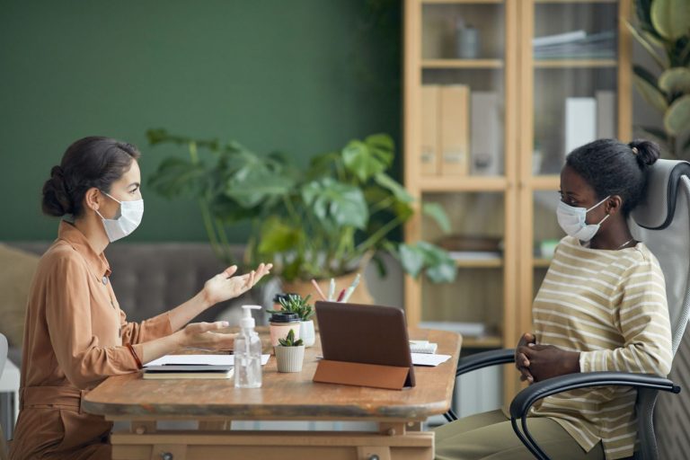 two women talking across a desk while wearing medical masks