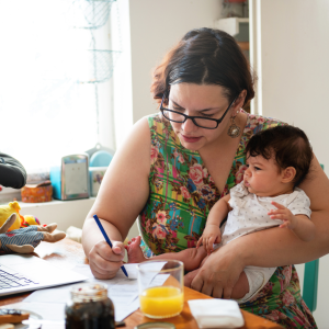 Woman sitting at computer on busy table, holding baby while writing